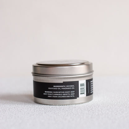 A small round metal tin candle with a silver lid and a black label, photographed on a light neutral surface against a pale background.