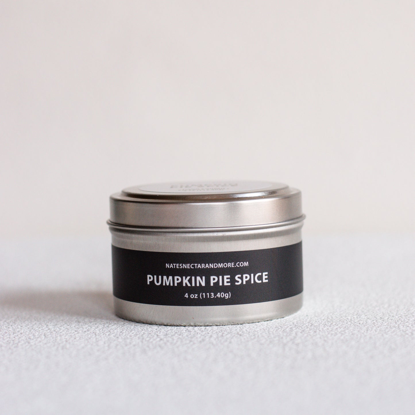 A small round metal tin candle labeled 'PUMPKIN PIE SPICE' by Natescigarandmore.com, with a black label and silver lid, photographed on a light neutral surface against a pale background.
