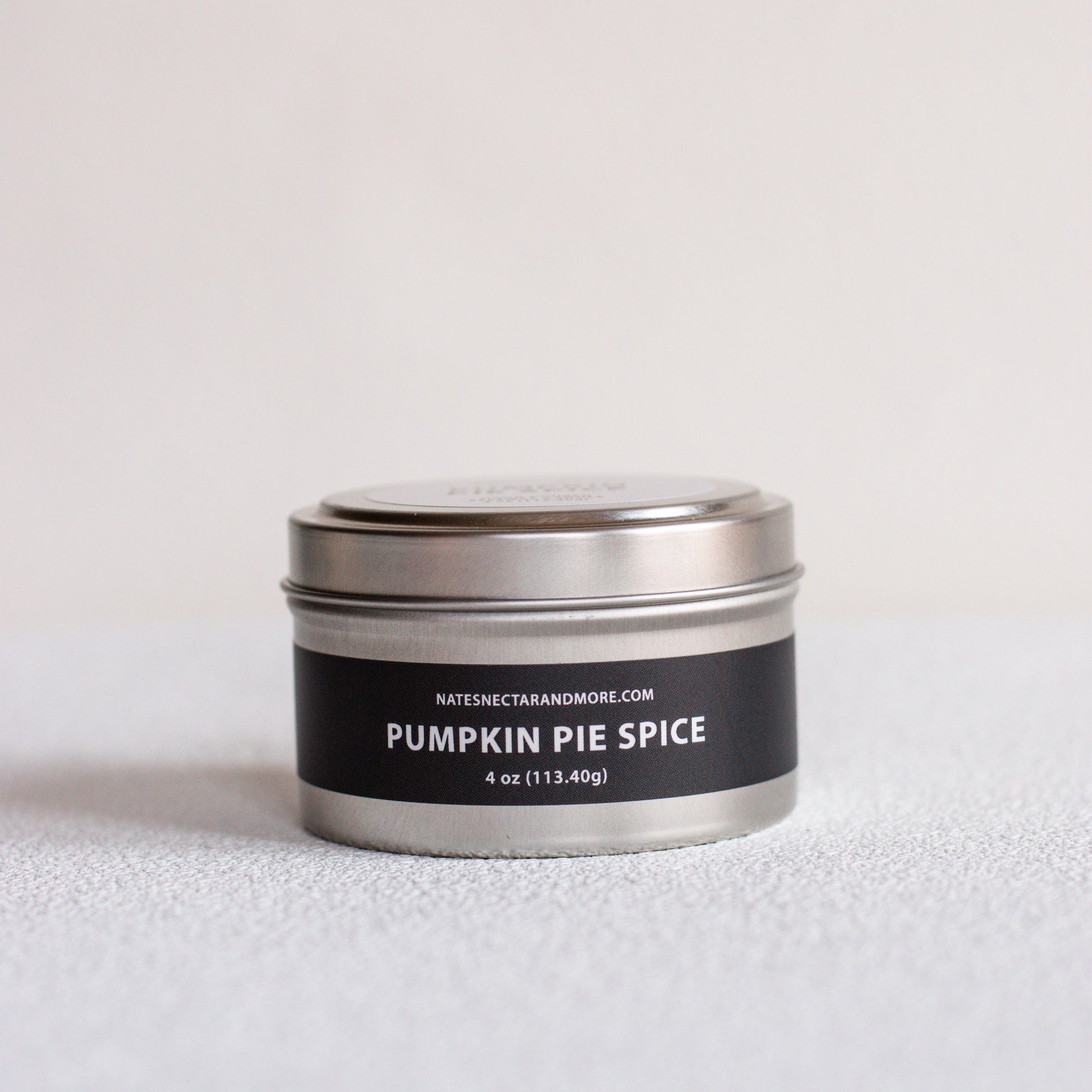 A small round metal tin candle labeled 'PUMPKIN PIE SPICE' by Natescigarandmore.com, with a black label and silver lid, photographed on a light neutral surface against a pale background.