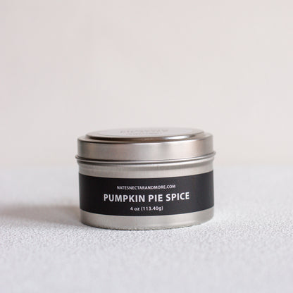 A small round metal tin candle labeled 'PUMPKIN PIE SPICE' by Natescigarandmore.com, with a black label and silver lid, photographed on a light neutral surface against a pale background.