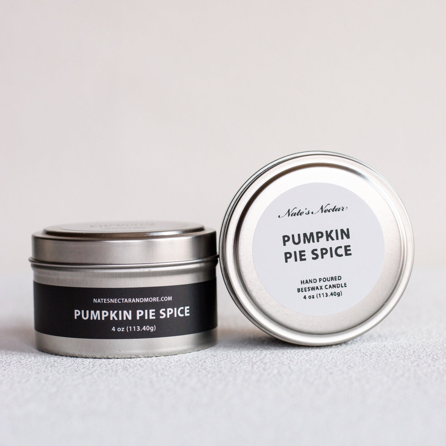 Two silver metal tins of Nates Nectar Candle in the Pumpkin Pie Spice scent, one closed with a black label and one open showing the white lid label, on a light gray textured surface.