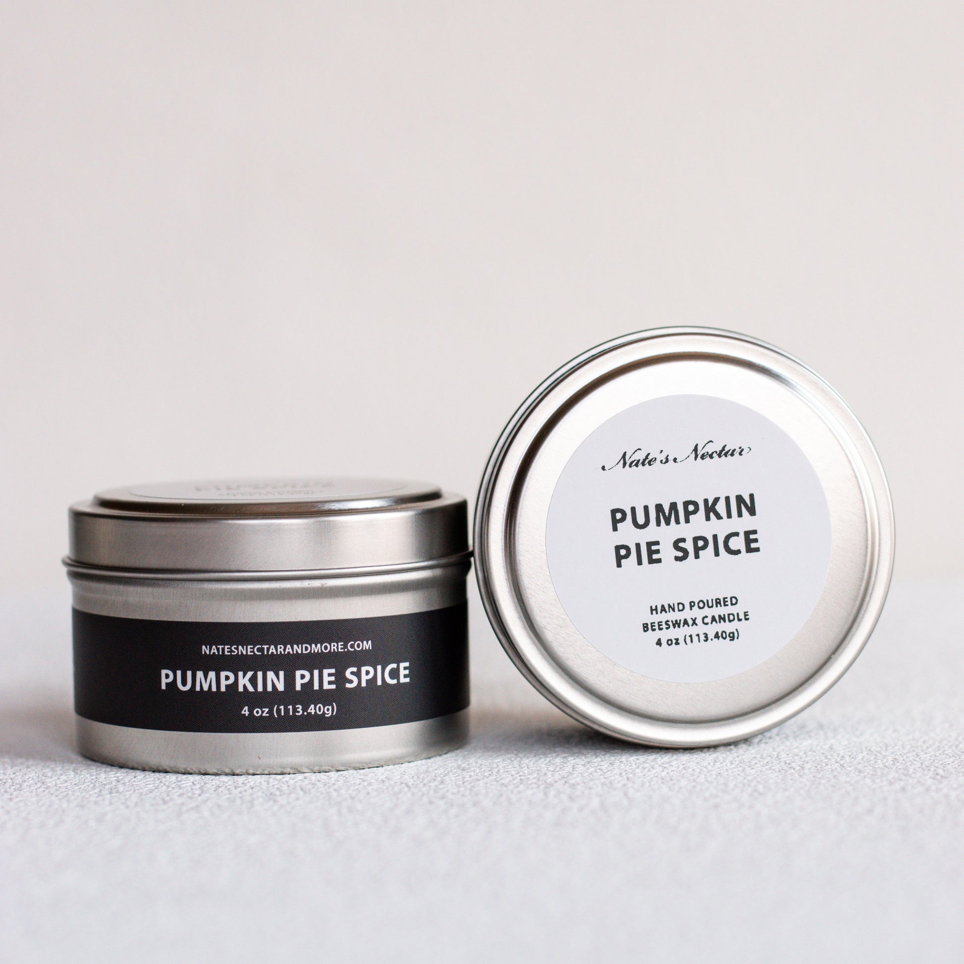 Two silver metal tins of Nates Nectar Candle in the Pumpkin Pie Spice scent, one closed with a black label and one open showing the white lid label, on a light gray textured surface.