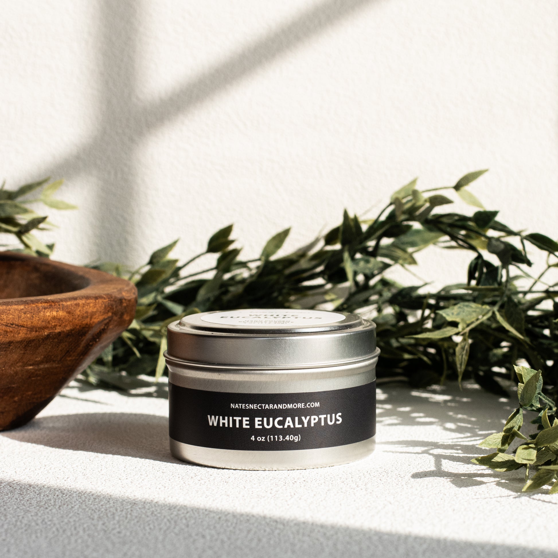 Candle tin labeled 'White Eucalyptus' on a surface with greenery in the background