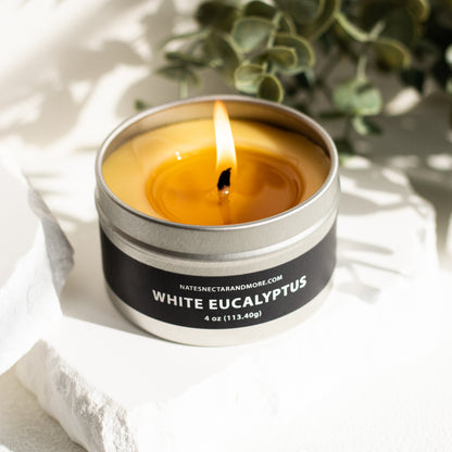 Candle in a metal tin labeled 'White Eucalyptus' on a light fabric background with greenery.